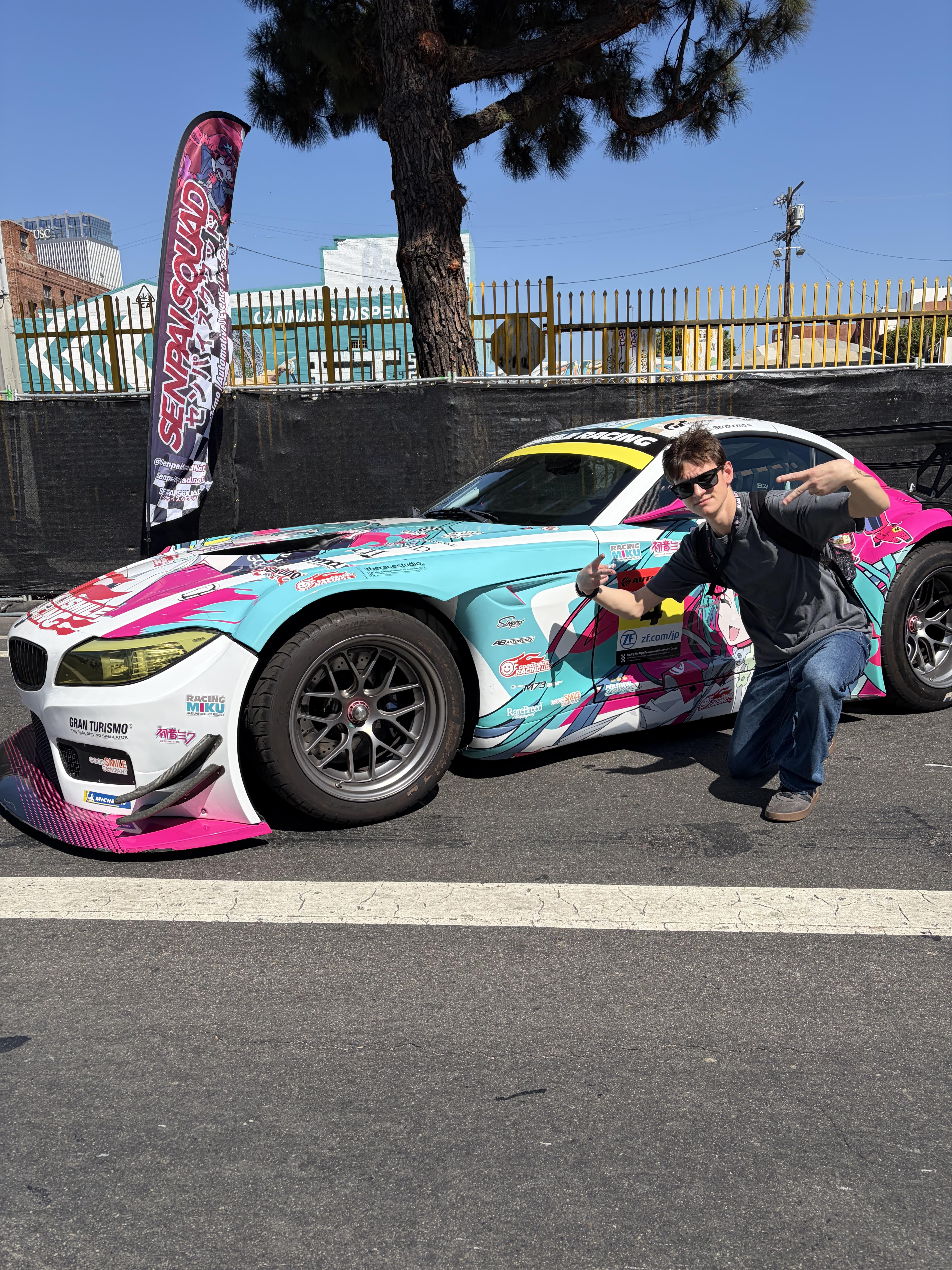 A photo of me standing in front of a custom-wrapped car at Anime Expo 2026 in Los Angeles, California. The car is small, but sporty, has two seats, the color scheme is white, blue, and pink, and there is a large flaming logo on the hood that says Good Smile racing in large italics. I am wearing a grey t-shirt and dark blue jeans.